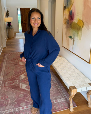 A woman wearing a matching navy blue long-sleeve top and wide-leg pants set stands in a home hallway with hardwood floors, a patterned rug, a woven bench, and a large abstract painting on the wall.