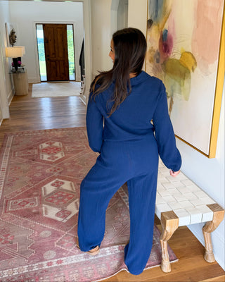 A woman wearing a royal blue long-sleeve jumpsuit stands in a hallway with hardwood floors, a patterned rug, and a large abstract painting on the wall.