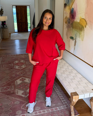 A woman modeling a matching red lounge set consisting of a short-sleeve top and sweatpants, standing in a home hallway next to a woven bench and a large abstract painting.