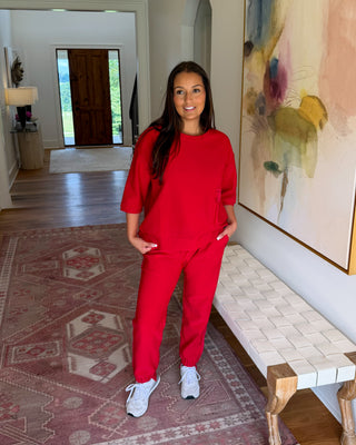 A woman wearing a matching red lounge set consisting of a short-sleeve top and sweatpants, standing in a home hallway next to a woven bench and a large abstract painting.
