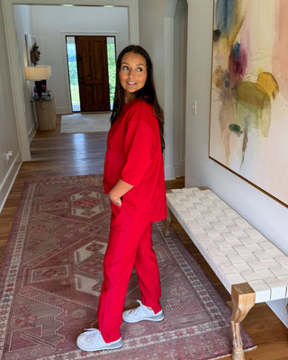 A woman wearing a bright red scrub set and white sneakers stands in a hallway, looking back over her shoulder. The hallway features a patterned rug, a woven bench, and a large abstract painting on the wall.