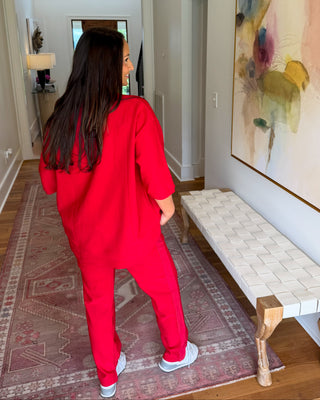 A woman wearing a bright red matching lounge set stands in a hallway, viewed from behind. The set appears to be a loose sweatshirt and jogger pants, with white sneakers.