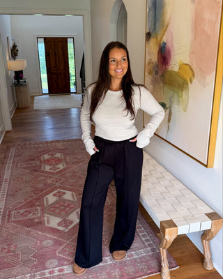 A woman stands in a home hallway wearing a white ribbed long-sleeve top and black wide-leg trousers, with a patterned rug, a bench, and a large abstract painting in the background.