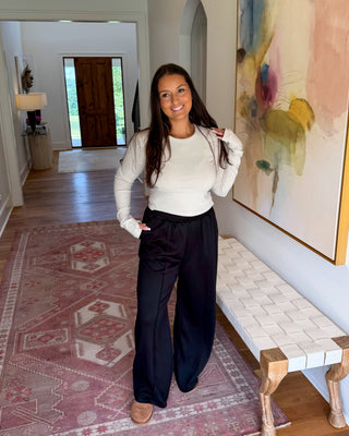 A woman stands in a home hallway wearing a white long-sleeve knit top and black wide-leg pants, with a patterned rug, a bench, and a large abstract painting in the background.