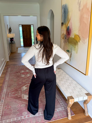 A woman stands in a hallway wearing a fitted white long-sleeve top and loose black wide-leg pants, photographed from the back. The setting includes a patterned rug, a bench, and a large abstract painting on the wall.