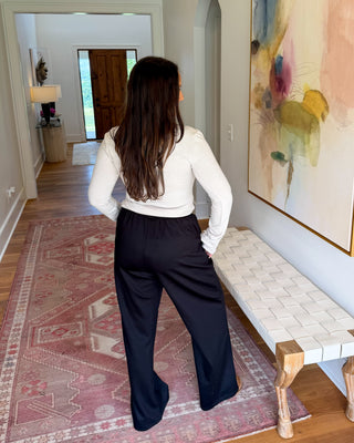 A woman stands in a hallway wearing a white long-sleeve top and black wide-leg pants, facing away from the camera. The setting features a patterned pink rug, a white bench with wooden legs, and a large abstract painting on the wall.