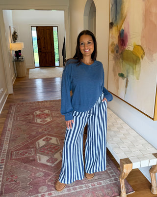 A woman stands in a home hallway wearing a blue long-sleeve top and blue-and-white vertically striped wide-leg pants. She is positioned next to a white woven bench and a large abstract painting on the wall, with a patterned rug and wooden floor visible.
