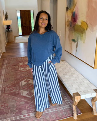 A woman stands in a home hallway wearing a dusty blue long-sleeve top and blue-and-white vertically striped wide-leg pants. She is positioned next to a woven bench and a large abstract painting, with a patterned rug and wooden floors visible.