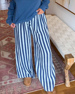 A woman wearing a blue long-sleeve top and blue-and-white vertically striped wide-leg pants stands in a home hallway with hardwood floors, a patterned rug, a bench, and a large abstract painting on the wall.