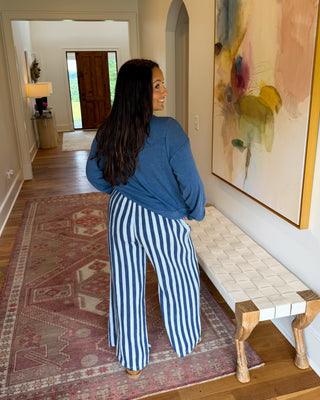 A woman stands in a hallway wearing a blue long-sleeve top and blue-and-white vertically striped wide-leg pants, looking to the side. A patterned rug, a bench, and a large abstract painting are visible in the room.