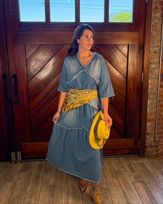 A woman stands in front of a large wooden door wearing a blue denim dress with a yellow patterned sash and holding a yellow hat. She is also wearing brown boots.