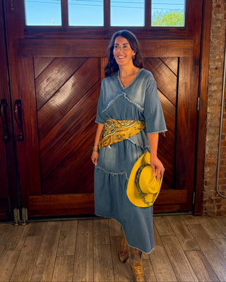 A woman stands in front of a large wooden door wearing a blue denim-style dress with a yellow patterned belt and holding a yellow hat. The photo is taken indoors on a wood floor with brick wall visible on the right.