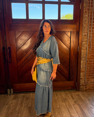 A woman stands indoors in front of a large wooden door wearing a light blue denim maxi dress with a yellow patterned belt and holding a yellow clutch.