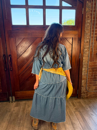 A woman stands with her back to the camera in front of a large wooden door, wearing a light blue denim dress with a yellow bandana tied around her waist and holding a yellow hat.