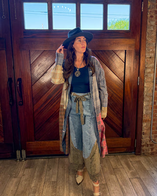 A woman stands in front of a large wooden door wearing a black wide-brim hat, a black top, a long plaid cardigan, a tie-waist denim skirt, and beige pointed-toe heels.