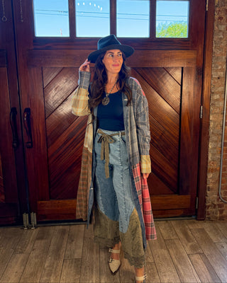 A woman stands in front of a large wooden door wearing a bohemian-style outfit featuring a black wide-brim hat, a black top, a long gray plaid coat, a blue denim skirt, and beige shoes.