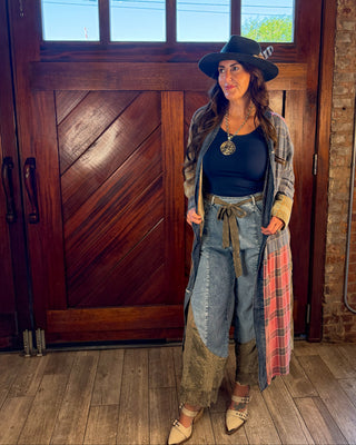 A woman stands in front of a large wooden door wearing a western-style outfit with a black wide-brim hat, black top, patchwork denim pants, a plaid cardigan, and pointed beige heels.