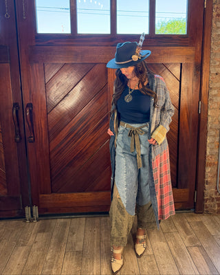 A woman stands in front of a large wooden door wearing a bohemian-style outfit featuring a blue hat with a feather, a black top, a plaid cardigan, high-waisted patchwork jeans, and pointed beige heels.