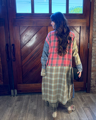 A woman stands with her back to the camera in front of a large rustic wooden door, wearing a patchwork plaid tunic dress with red, green, and beige tones over layered blue and yellow shirts.