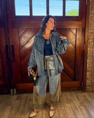 A woman stands in front of a large wooden door wearing a distressed denim jacket, patchwork wide-leg jeans in blue and beige, a black top, and beige pointed-toe heels. She is holding a small black clutch.