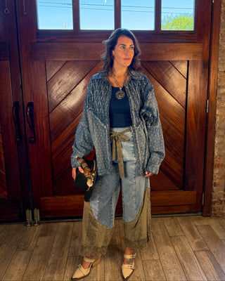 A woman stands in front of a large wooden door wearing a layered outfit consisting of a distressed denim jacket, a dark navy top, light-wash patchwork wide-leg jeans with a tan tie belt, and beige pointed-toe heels. She is holding a small black handbag.