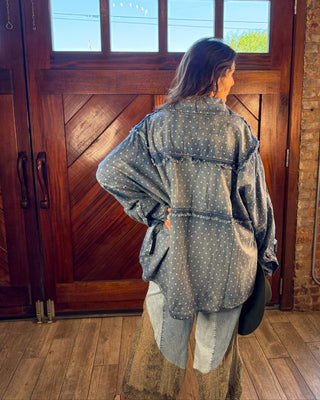 A woman stands with her back to the camera in front of a large wooden door, wearing a loose gray denim jacket with white polka dots and light-wash denim bottoms.