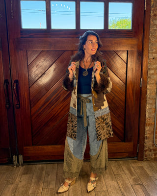 A woman stands in front of a large wooden door wearing a bohemian-style patchwork coat with animal print and denim details, a dark blue top, and light-colored pointed shoes.