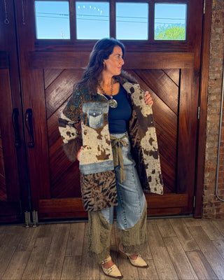 A woman stands in front of a large wooden door wearing a bohemian-style patchwork coat with animal print, denim pocket details, and faux fur trim, paired with a dark top, light-wash jeans, and tan shoes.