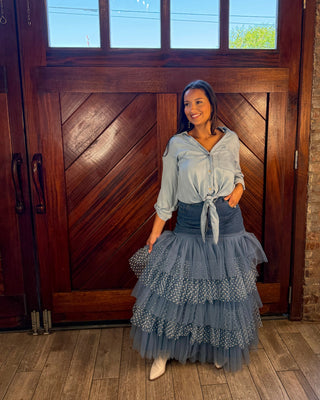 A woman stands in front of a large wooden door wearing a light blue tie-front blouse and a long, tiered dusty blue tulle skirt with white polka dots. She is holding a dark clutch and wearing white pointed shoes.