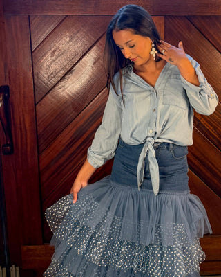 A woman posing in front of a rustic wooden door, wearing a light blue long-sleeve button-up shirt tied at the waist and a layered, tiered slate blue tulle skirt with white polka dots.