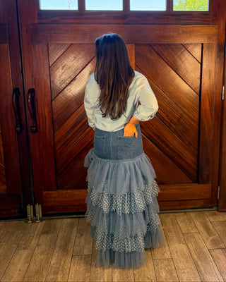 A woman viewed from behind stands in front of a large wooden door wearing a white long-sleeve top, a denim waistband, and a layered gray tulle skirt with white patterned trim.