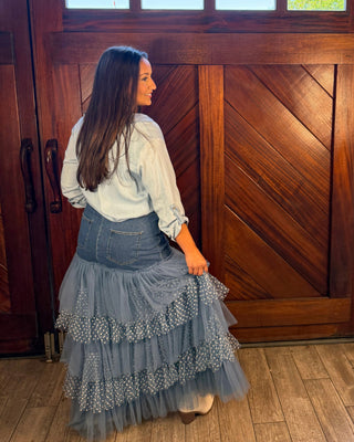 A woman stands in front of a large rustic wooden door, wearing a light blue long-sleeve blouse and a layered gray tulle skirt with small white polka dots.