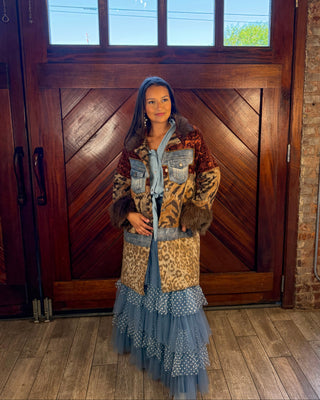 A woman stands in front of a large wooden door wearing a long, layered fashion outfit with a leopard-print jacket, denim vest, blue tulle skirt, and a blue head covering.