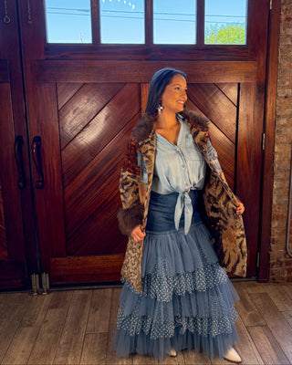 A woman stands in front of a large wooden door wearing a layered outfit featuring a leopard-print coat with brown fur trim, a light blue tie-front blouse, a dark blue corset-style waist, and a tiered blue tulle skirt.