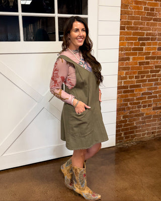 A woman posing in a rustic barn setting, wearing an olive green sleeveless apron over a pink patterned long-sleeve shirt and distressed brown cowboy boots.