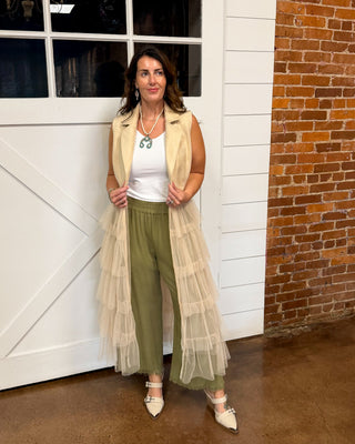 A woman stands in front of a white barn-style garage door and exposed brick wall, wearing a long cream tulle vest over a white top and olive green wide-leg pants with white pointed flats.