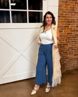 A woman modeling a sleeveless beige tulle vest over a white top and blue wide-leg pants, standing in front of a white barn-style garage door and brick wall.