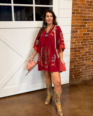 A woman stands in front of a white barn-style garage door and brick wall, wearing a loose red bohemian-style mini dress with colorful paisley and floral patterns, tan cowboy boots, and holding a pink handbag.