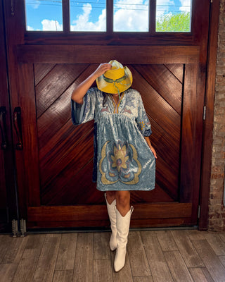 A woman stands in front of a large wooden door wearing a light blue patterned dress, a yellow hat, and white cowboy boots. The photo is taken indoors with a visible brick wall and wood floor, and the person's face is obscured by the hat.