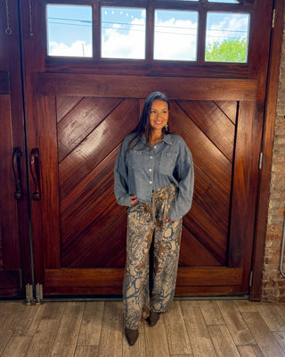 A woman stands in front of a large wooden door wearing a light blue denim shirt and high-waisted, wide-leg pants with a brown and beige snakeskin pattern. She is wearing brown boots.