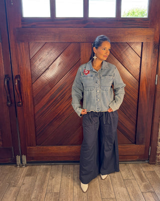 A woman stands in front of a large wooden door wearing a light blue denim jacket with a red patch on the chest, dark wide-leg pants, and white pointed shoes.