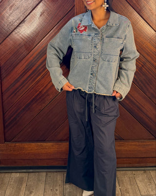 A woman stands in front of a large wooden door wearing a light-wash denim jacket, dark wide-leg pants, and white pointed shoes.