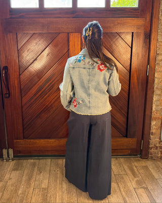 A woman viewed from behind stands in front of large wooden double doors, wearing a light blue denim jacket with colorful floral embroidery on the back and dark wide-leg pants.