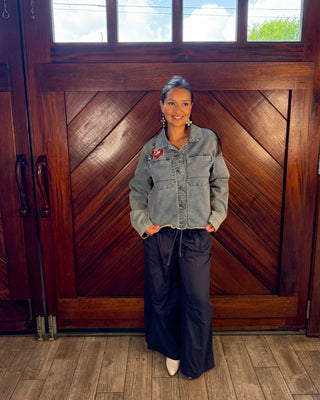 A woman stands in front of a large wooden door wearing a light-wash denim jacket with a red embroidered patch on the chest, dark wide-leg pants, and white pointed shoes.
