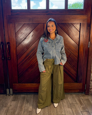 A woman stands in front of a large wooden door wearing a light blue cropped denim jacket, olive green wide-leg pants, and white pointed shoes.