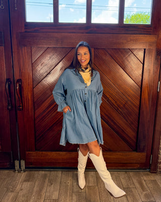 A woman stands in front of a large wooden door wearing a light blue denim-style dress with long sleeves, a gathered neckline, and front buttons, paired with a yellow top and off-white knee-high boots.
