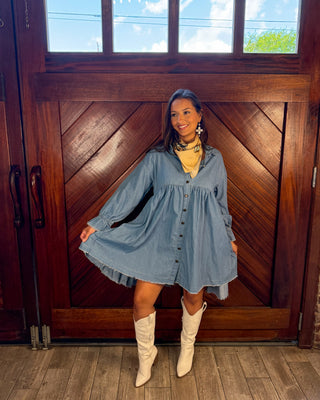 A woman stands in front of a large wooden door wearing a light blue denim shirt dress with a yellow top and white cowboy boots.