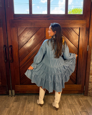A woman wearing a light blue tiered long-sleeve dress and white knee-high boots stands in front of a large wooden door, holding out the sides of the dress to show its full width.