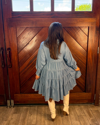 A woman viewed from behind wearing a light blue tiered long-sleeve dress and cream-colored boots, standing in front of a large rustic wooden door.