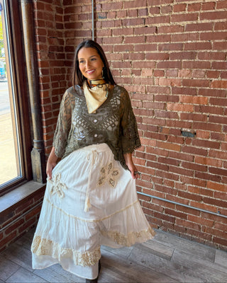 A woman wearing a bohemian-style outfit consisting of a brown lace-trimmed top, a cream neck scarf, and a long white tiered skirt with gold embroidery, posing in front of a red brick wall.
